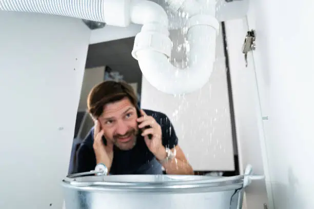 Stressed man on the phone looking up at a severely leaking white PVC P-trap under a sink, with water pouring into a metal bucket below in what appears to be an older house.