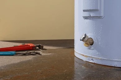 Close-up of a white water heater unit drain spigot with a red pipe wrench lying on the concrete floor.