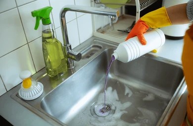 Gloved hands pouring purple liquid chemical drain cleaner into a stainless steel kitchen sink drain.