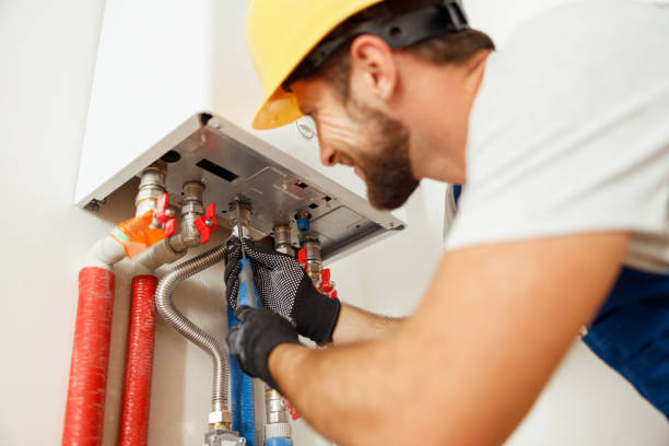 Plumber in a hard hat using tools to repair pipe connections under a wall-mounted water heater.