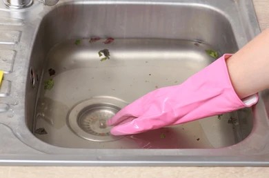Hand wearing a pink rubber glove reaching into a clogged kitchen sink filled with dirty standing water and food debris.