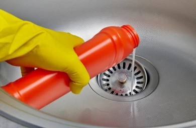 Hand wearing a yellow rubber glove pouring liquid chemical drain cleaner from an orange bottle into a sink drain.
