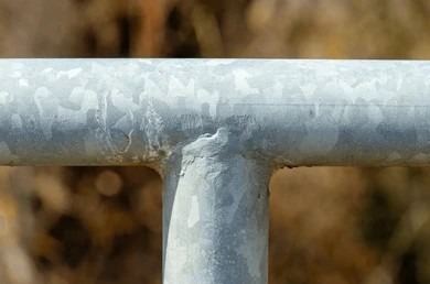 Close-up of dull gray galvanized steel pipe T-joint showing characteristic spangle pattern.